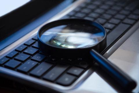Image of a magnifying glass resting on a laptop keyboard, with the glass magnifying a portion of the keys.