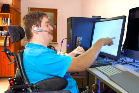 A person in a wheelchair wearing a blue shirt and headset, working at a desk with two computer monitors, pointing at one screen, in a room with bookshelves and a computer tower.